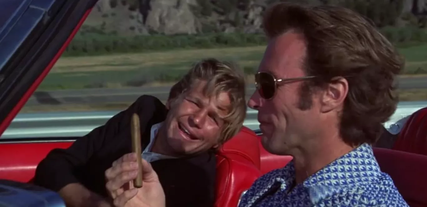 Thunderbolt hands Lightfoot a cigar as the two drive in a convertible in Thunderbolt and Lightfoot.