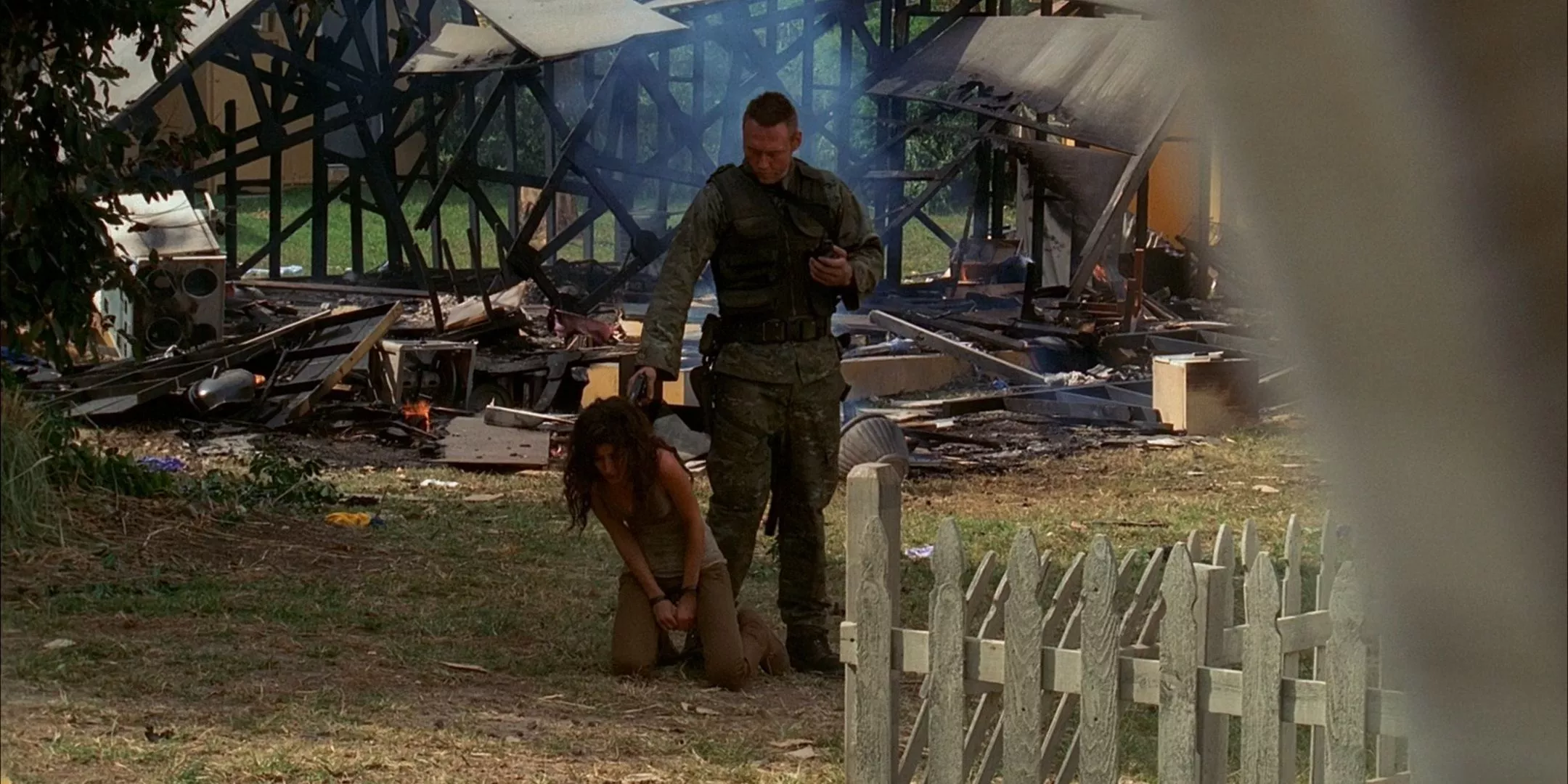 Keamy stands in front of a burned out house while a fearful alexandra kneels on the ground and he points a gun at her head from the Lost series