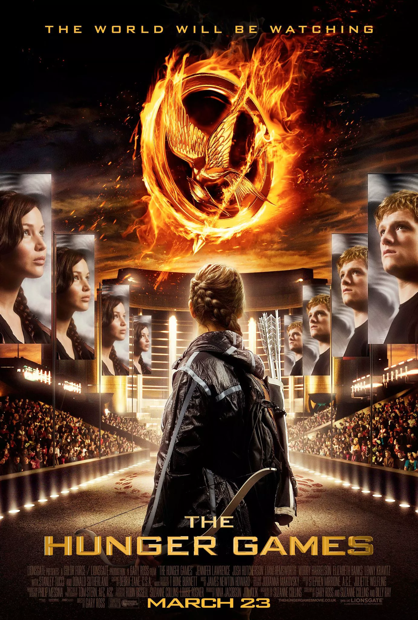 Katniss stands facing posters of her and Peeta with the Mockingjay symbol on fire in the center in The Hunger Games First Film Poster