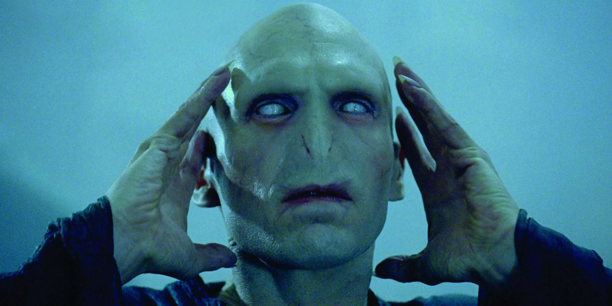 Ralph Fiennes como Voldemort con ojos blancos, sostiene sus manos a los lados de su cabeza en Harry Potter.