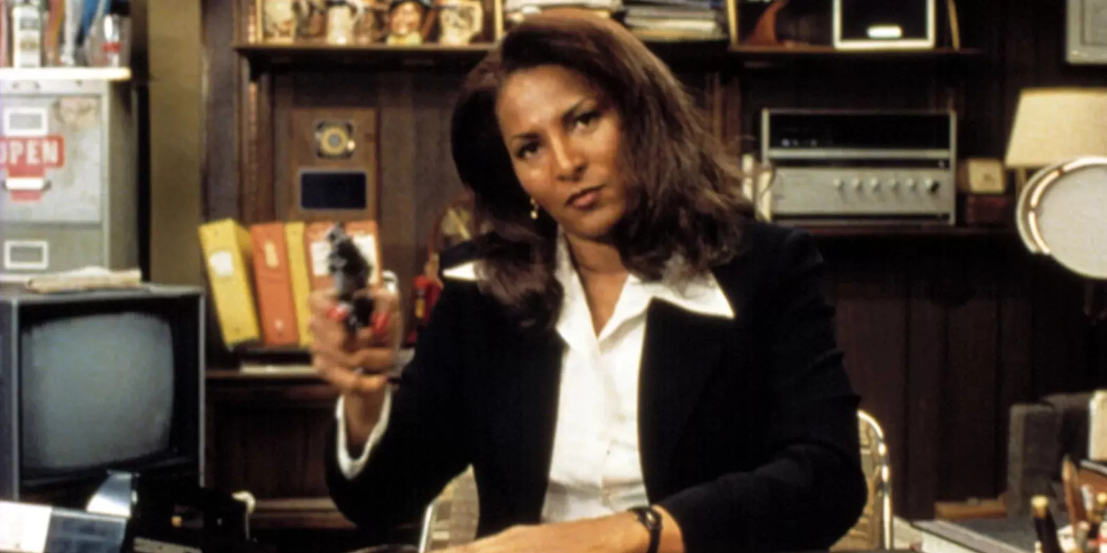 Pam Grier as Jackie Brown sits at a desk in a suit and points a pistol in Jackie Brown